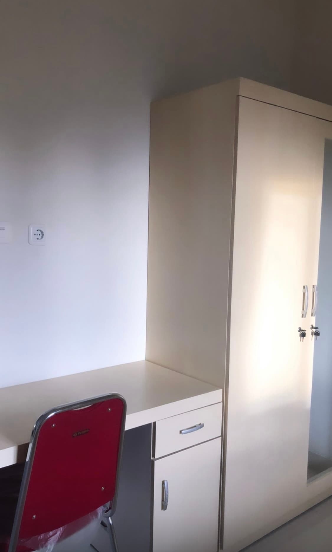 A minimalist workspace is presented with a light-colored desk and a red chair. Adjacent stands a tall wardrobe with a mirrored door, reflecting the room's bright ambiance. The neutral walls contribute to a calm and organized environment.