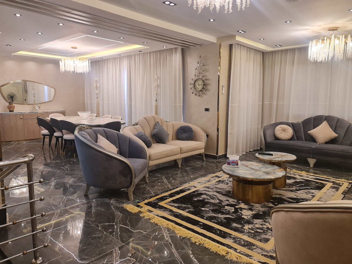 An elegantly furnished living area features two plush sofas, arranged around a coffee table with a marble top. Soft, neutral tones are complemented by a modern chandelier above. Large windows with sheer curtains allow natural light to brighten the space, enhancing the sophisticated decor.