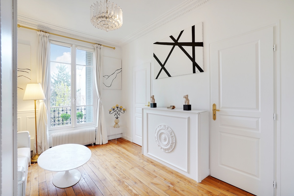 A bright living area features a large window that allows natural light to enter. Elegant white walls are adorned with abstract artwork. Light wooden flooring complements a round coffee table, and a chic lamp adds a touch of coziness. Decorative elements create a refined atmosphere.