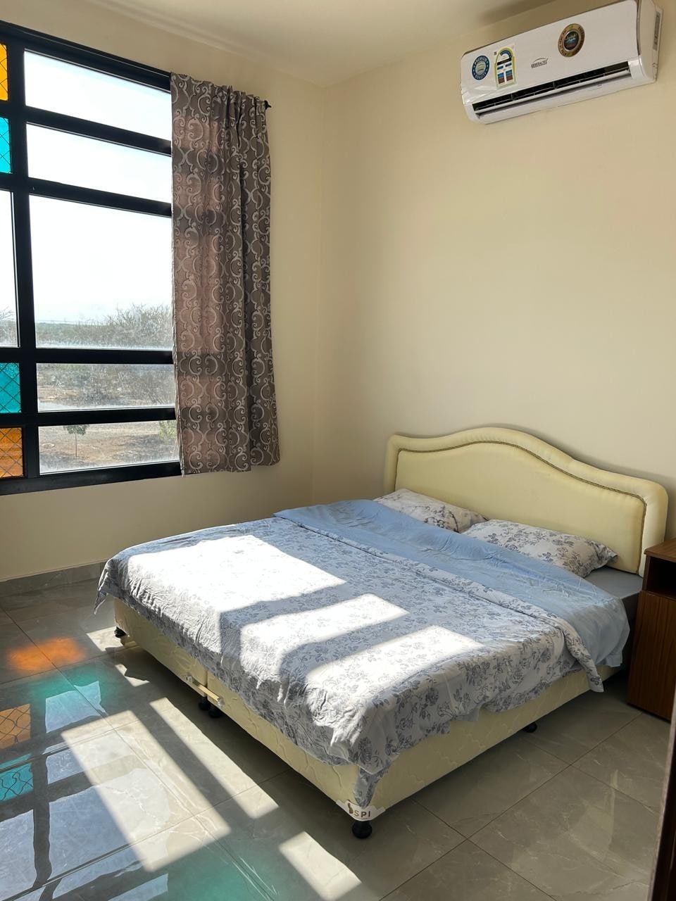 A bright sleeping area features a spacious bed with a light-colored bedspread. Natural light streams through large windows, casting shadows on the tiled floor. Soft curtains frame the windows, and a small wooden nightstand is positioned beside the bed.