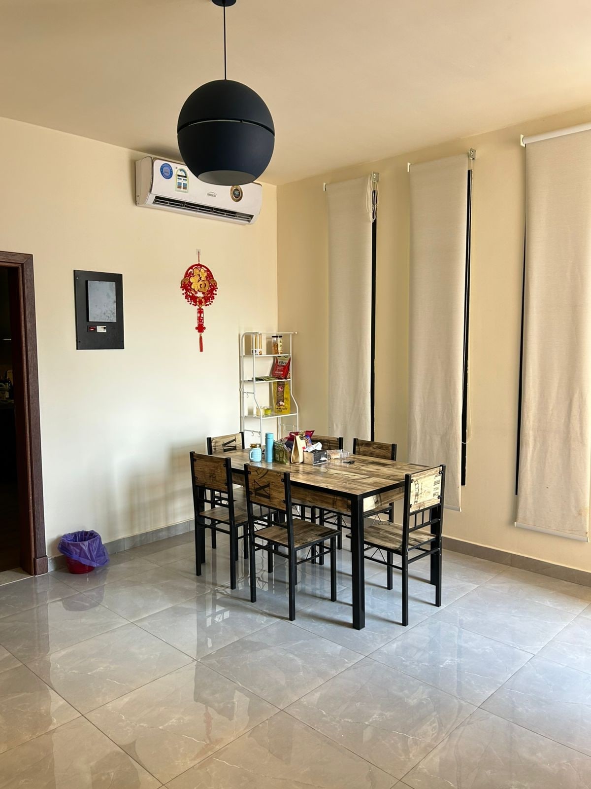 A dining area features a rectangular table surrounded by six black chairs. A glass display shelf is positioned against the wall, holding assorted items. Large windows are dressed with light-colored curtains, allowing natural light to brighten the space.