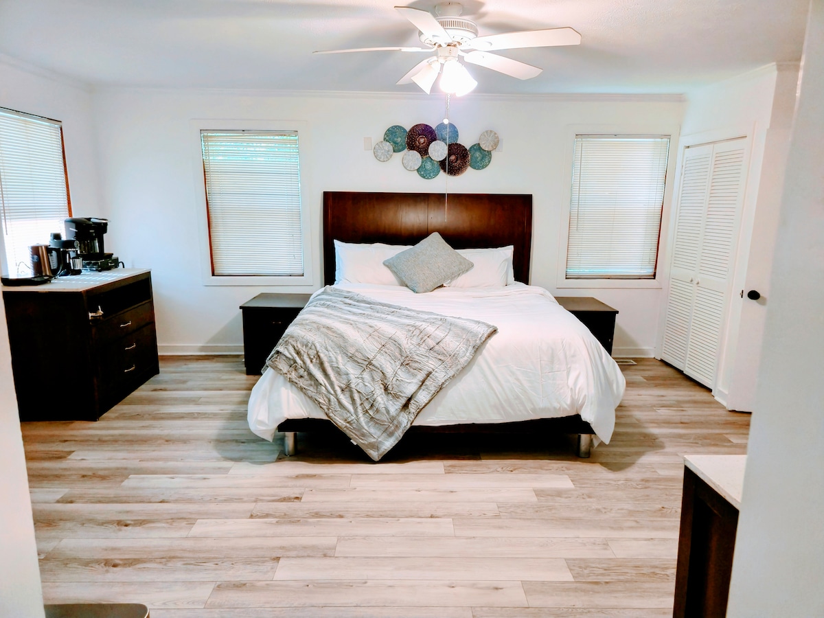 A spacious bedroom features a queen-sized bed with a decorative throw and a fan overhead. Nightstands are placed on either side, and a coffee station is visible against the wall. Natural light enters through two windows, with a closet in the background.