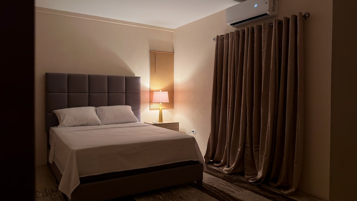 A cozy bedroom is presented with a spacious bed adorned with crisp white linens. Soft, warm light is emitted from a bedside lamp, illuminating the room's calming color palette. Heavy curtains frame the window, enhancing privacy and comfort, while air conditioning is visible above.