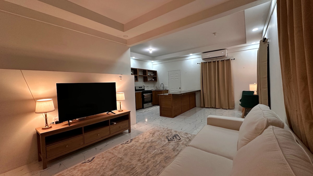 A spacious living area features a large television on a wooden media console, flanked by two table lamps. The open kitchen is visible in the background, accentuated by warm lighting. A comfortable sofa rests thoughtfully on the polished floor, inviting relaxation.