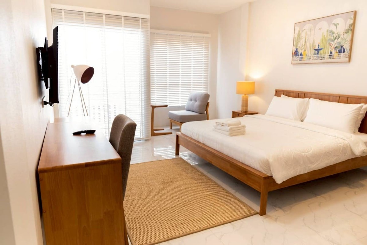 A bright and airy bedroom features a spacious bed covered with white linens. Natural light filters through blinds on large windows. A comfortable armchair sits beside a small side table, and a desk with a chair is positioned against the wall.