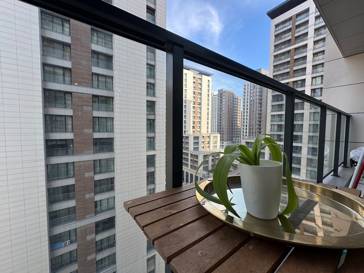 A balcony is featured with a wooden table supporting a small potted plant and a white mug on a circular tray. The view showcases an array of tall buildings under a clear blue sky, creating an urban setting.