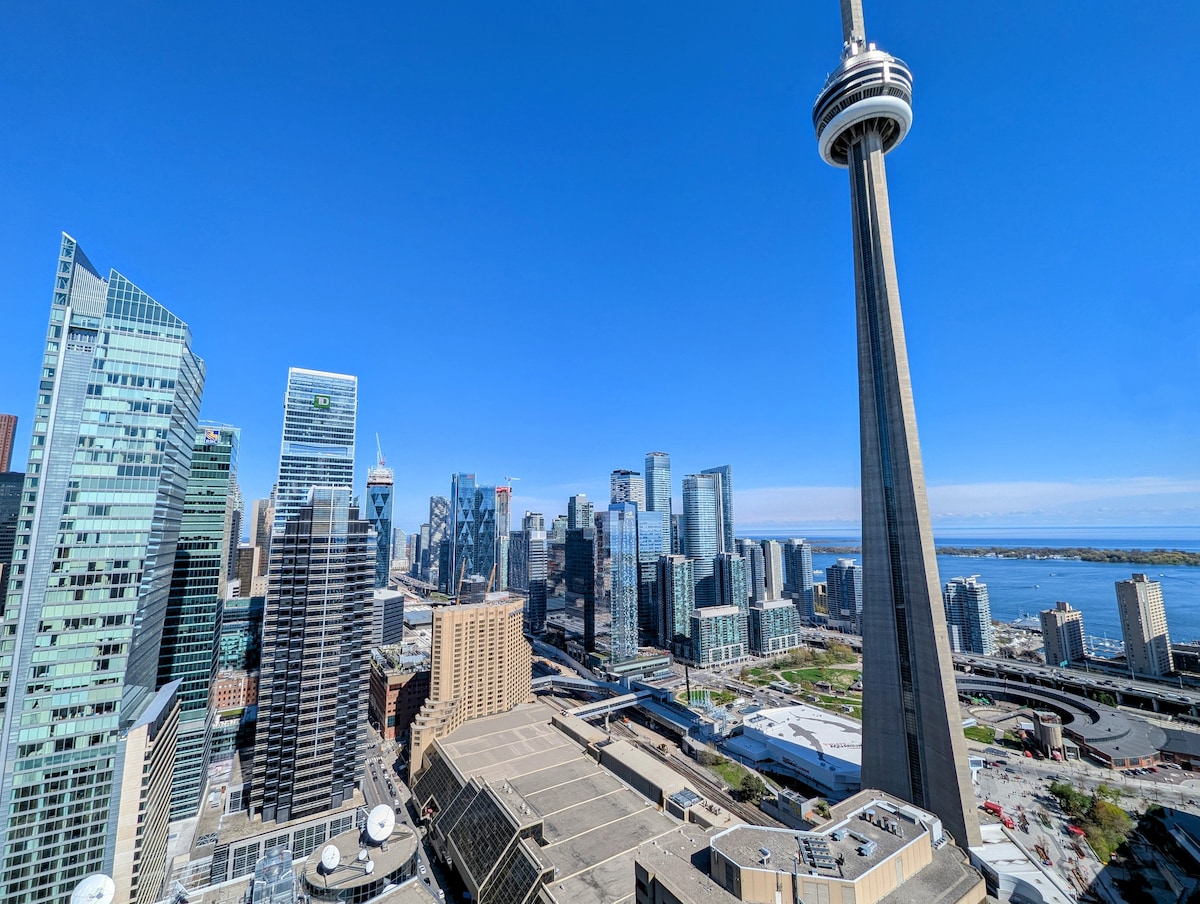 A panoramic view of Toronto's bustling skyline is showcased, featuring a prominent CN Tower alongside modern skyscrapers. The clear blue sky contrasts with the urban landscape, while Lake Ontario can be seen in the distance, contributing to the expansive cityscape.