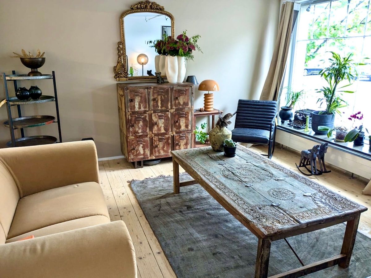 A cozy living area features a beige sofa and a rustic wooden coffee table, adorned with a decorative bowl and plants. A large mirror reflects natural light, while a black chair and a shelf with plants and decorative items complement the space.