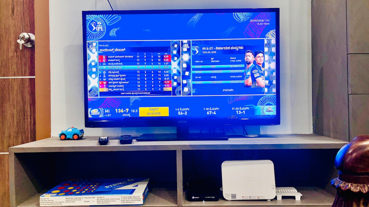 A 40-inch smart TV displays a live sports event, featuring scoreboards on both sides. Below the TV, a storage unit holds a collection of books and a small blue toy car. A wireless router is positioned on the right side.