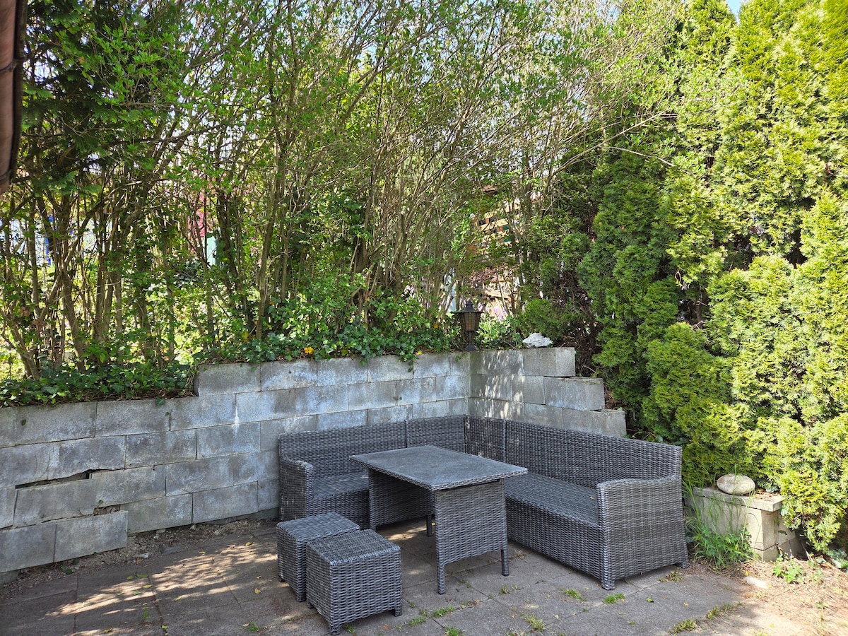 An outdoor seating area is presented, featuring a gray wicker sofa set and a matching coffee table. Surrounding greenery adds a natural backdrop, providing privacy. A stone wall supports the seating area, while a variety of plants contribute to the tranquil setting.