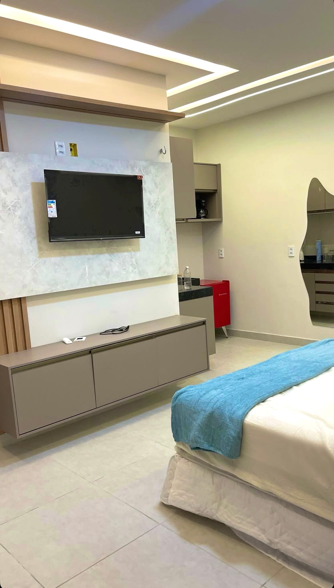 A modern room features a spacious bed with a white coverlet and blue accent blanket. A flat-screen TV is mounted on a light stone wall unit, complemented by a sleek console. Additional furnishings include a small kitchenette area with minimalist cabinetry.