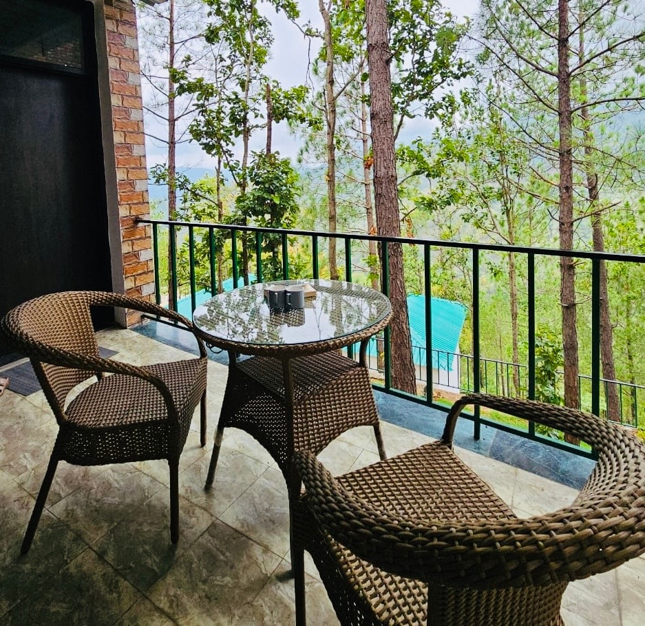 A small balcony area is featured, furnished with a round glass table and two wicker chairs. Lush pine trees are visible in the background, with scenic views of the mountain valley enhancing the natural setting. A hint of the resort's pool can be seen in the distance.
