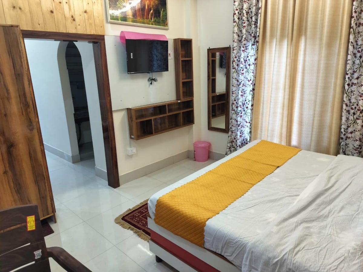 A calm bedroom space is fitted with a spacious bed, draped in a light-colored sheet with a vibrant yellow blanket. Floral-patterned curtains frame a window that allows natural light to fill the room. A wall-mounted television and wooden shelves offer functional features.
