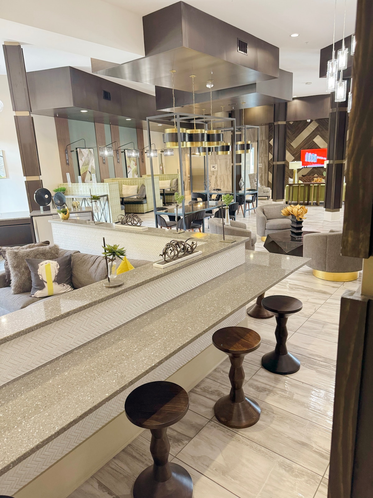 An elegant lobby area features a shimmering counter with contemporary bar stools arranged in front. Cozy seating is visible in the background, surrounded by stylish decor and large windows that invite natural light into the space.