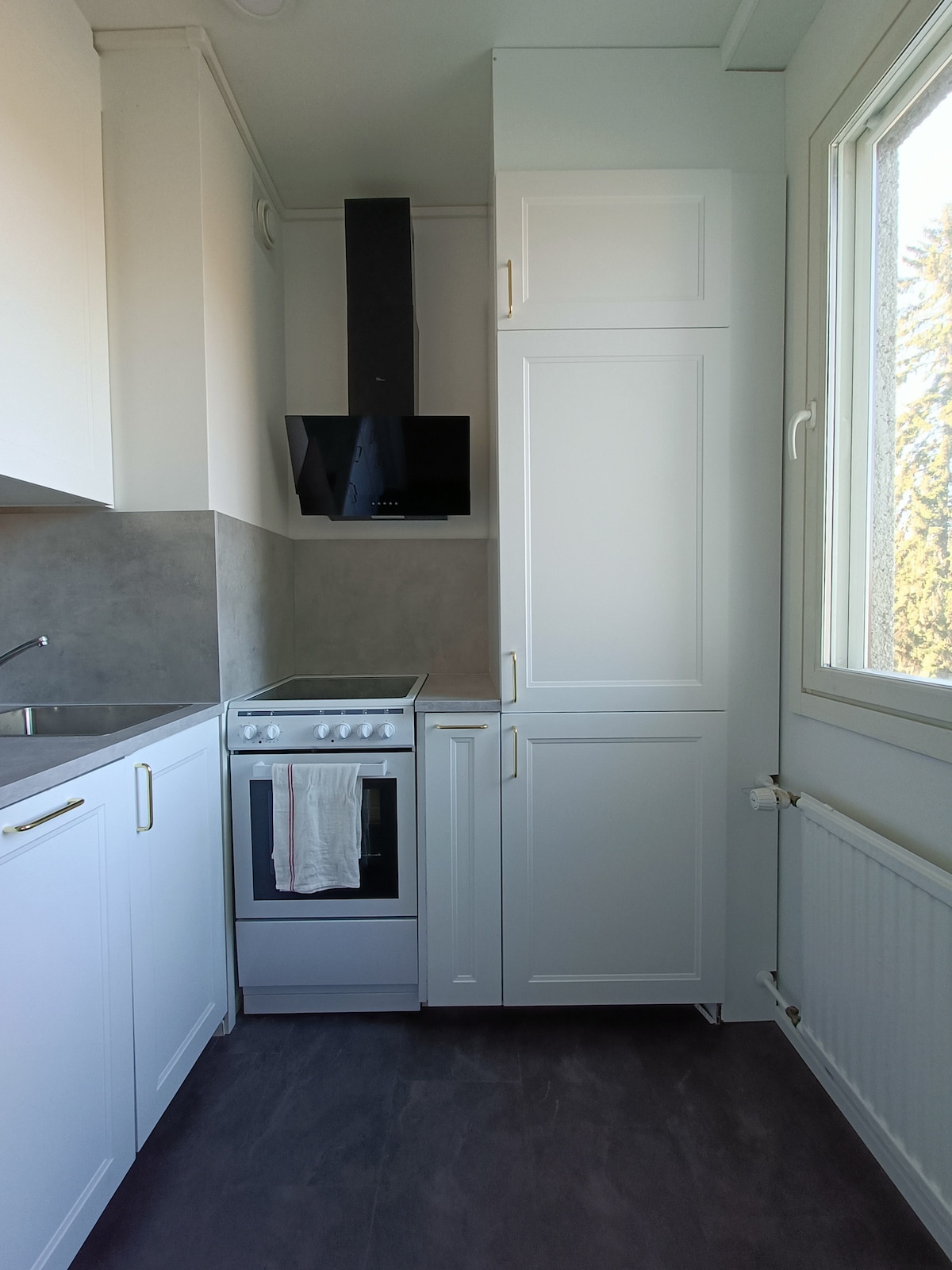 A modern kitchen area is presented with white cabinetry and gray countertops. An oven and stovetop are visible, with a chimney hood mounted above. Natural light enters through a window, highlighting the spacious and functional layout.