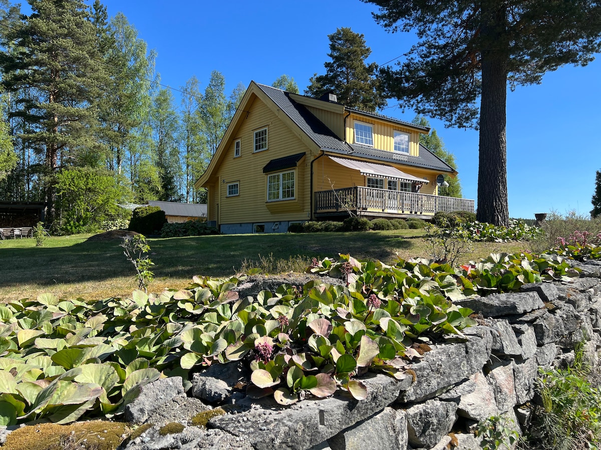The spacious yellow house is set amidst lush greenery, surrounded by trees. A stone wall with flowering plants lines the front, leading up to a covered porch with seating. The clear blue sky offers a bright backdrop to the inviting landscape.