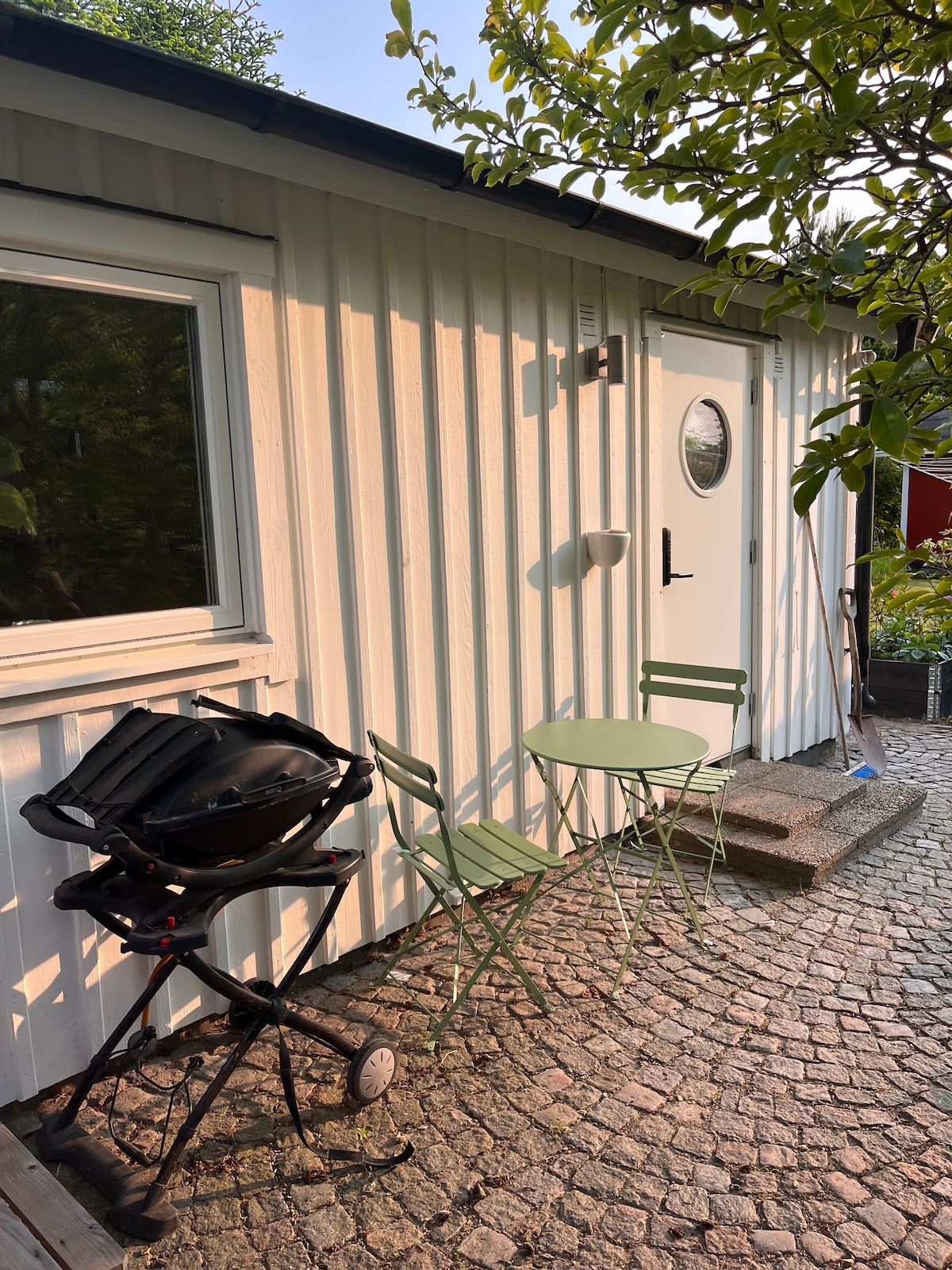 A stone-paved outdoor area features a compact circular table with two green folding chairs, positioned next to a gas grill. The entrance of the guest house is visible, characterized by a circular window and a simple door, enhancing the charming exterior.
