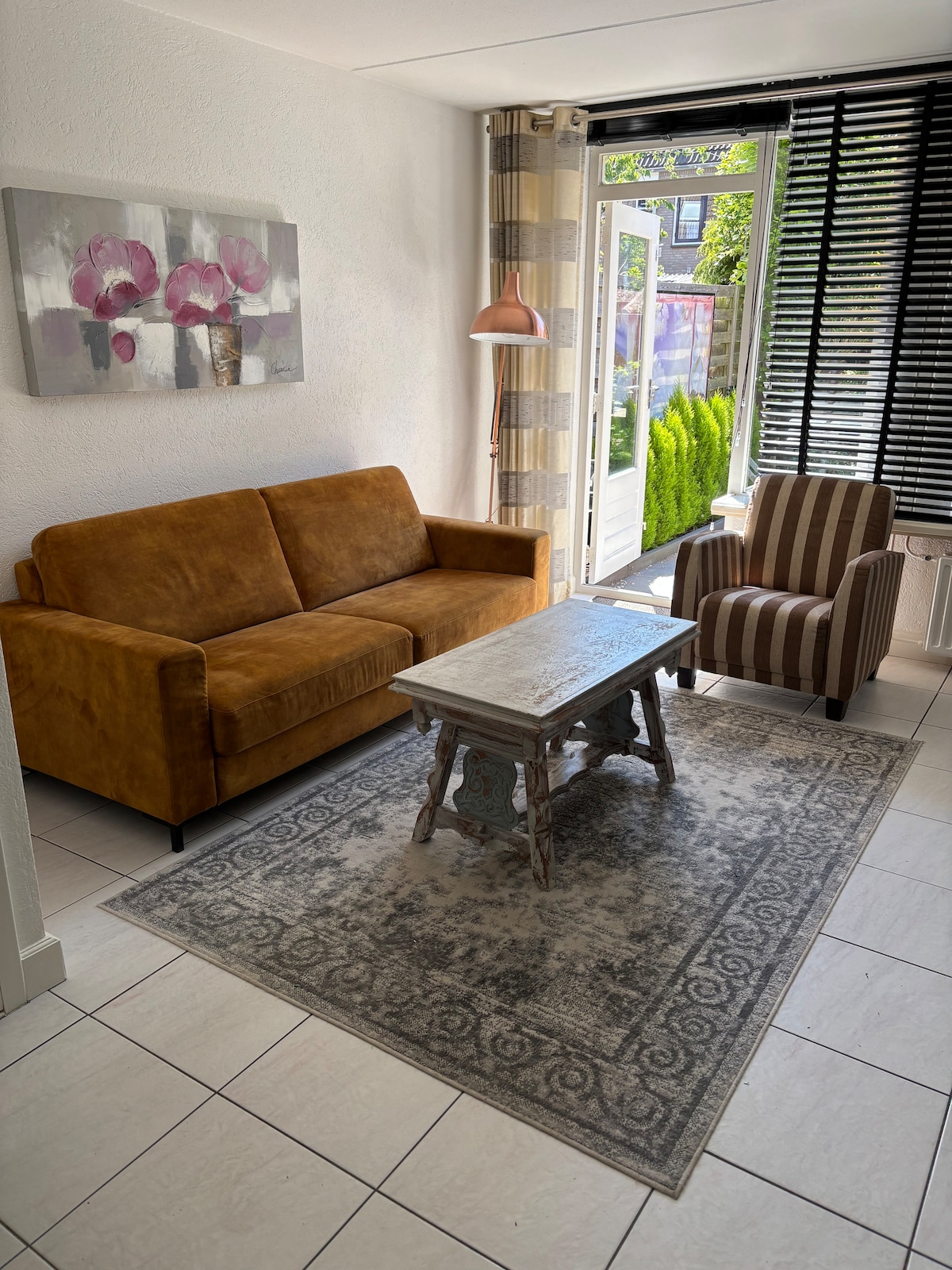 A cozy living area features a brown sofa and a striped accent chair, positioned around a weathered wooden coffee table. Natural light enters through glass doors, illuminating the space, while a piece of art with floral designs adorns the wall. A lush outdoor area is visible beyond the doors.