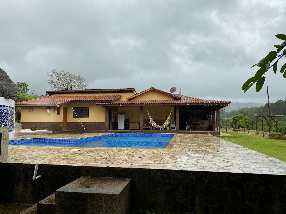 A spacious outdoor area is displayed, featuring a rectangular swimming pool surrounded by a stone deck. A charming house with a terracotta roof stands nearby, complemented by a hammock and lush green landscape in the background. Overcast skies are visible above.
