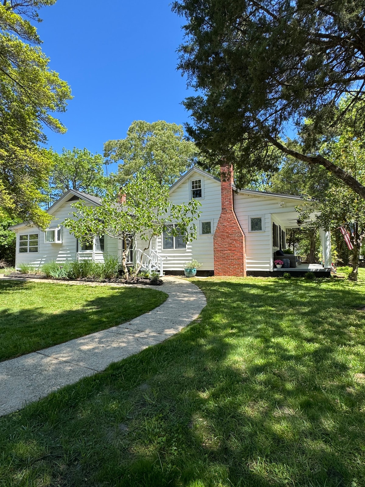 A charming bungalow is set against a clear blue sky, featuring a classic white exterior surrounded by lush greenery. A pathway leads to the entrance, where a red brick chimney adds a touch of color to the inviting facade.