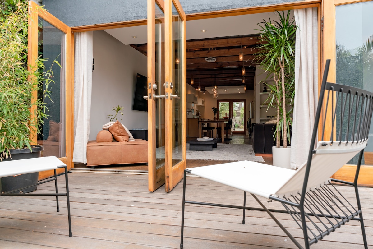 An inviting outdoor deck features two contemporary chairs set against wooden flooring, seamlessly connecting to the interior living space. Large French doors are open, allowing natural light to flood the room beyond, where designer furnishings and greenery create a relaxed atmosphere.