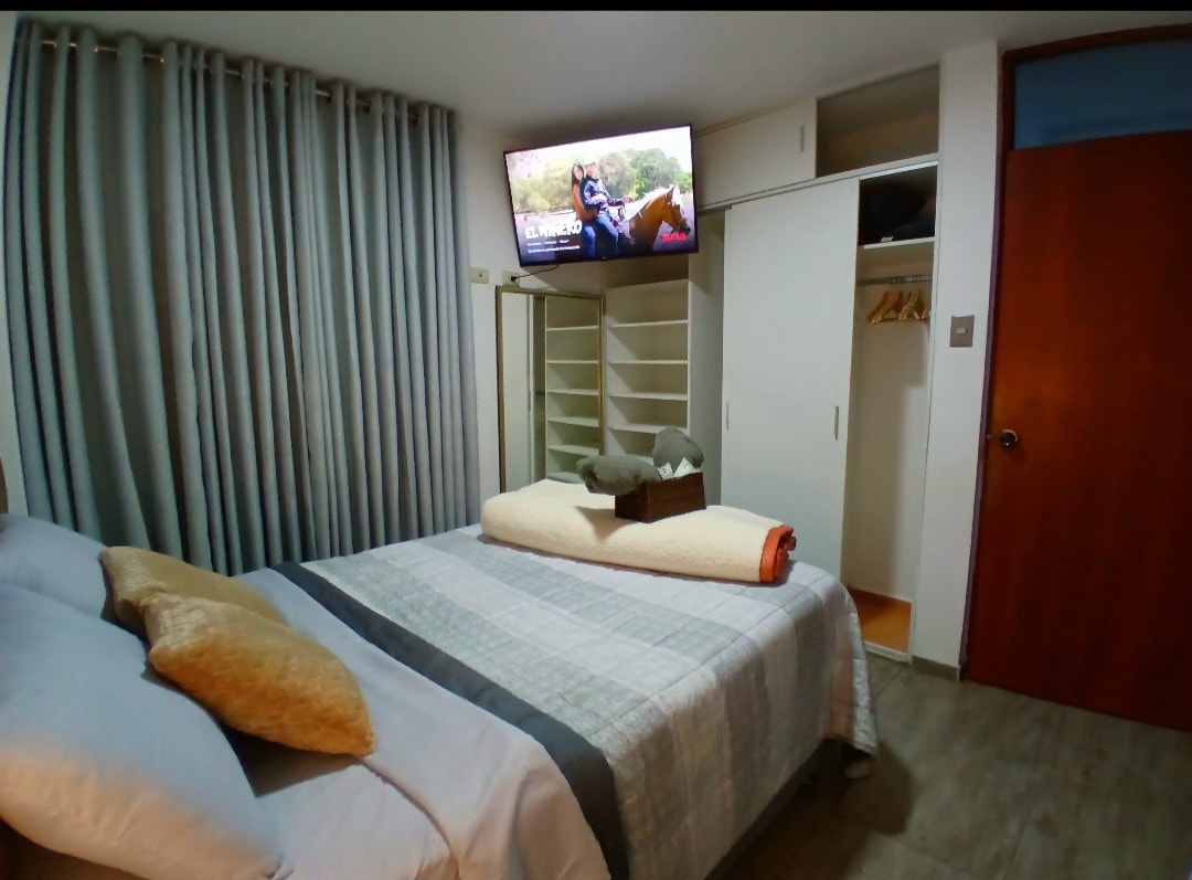 A modern bedroom features a spacious bed topped with a soft blanket and decorative pillows. A wall-mounted television is positioned across from the bed. A closet with shelves and hanging space is visible, complemented by thick curtains blocking natural light.