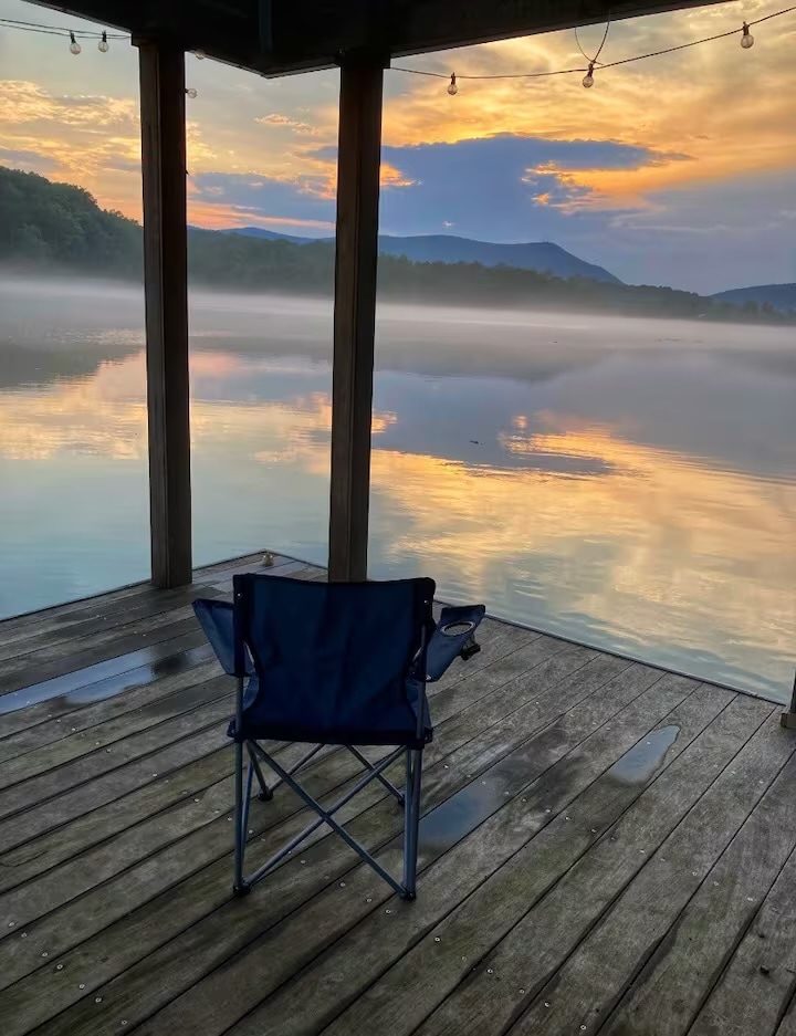 A lone chair is positioned on a wooden dock overlooking a serene lake. Soft morning light reflects off the water, with mist rising from the surface. Rolling mountains are visible in the background, enveloped in a calming orange and blue sky.