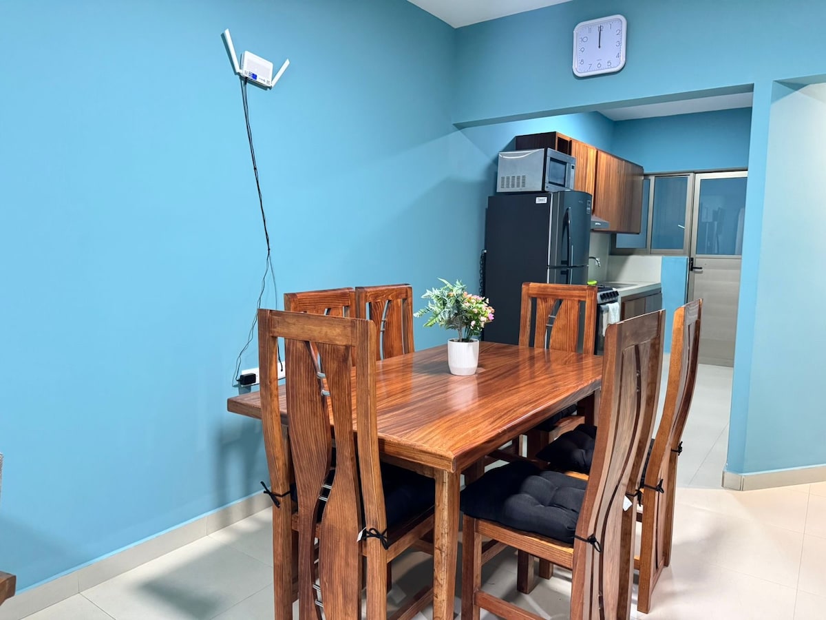 An inviting dining area is highlighted by a wooden table surrounded by six chairs. A potted plant sits at the center of the table. The walls are painted a calm blue, and a clock is mounted above, while a sleek kitchen is visible in the background.