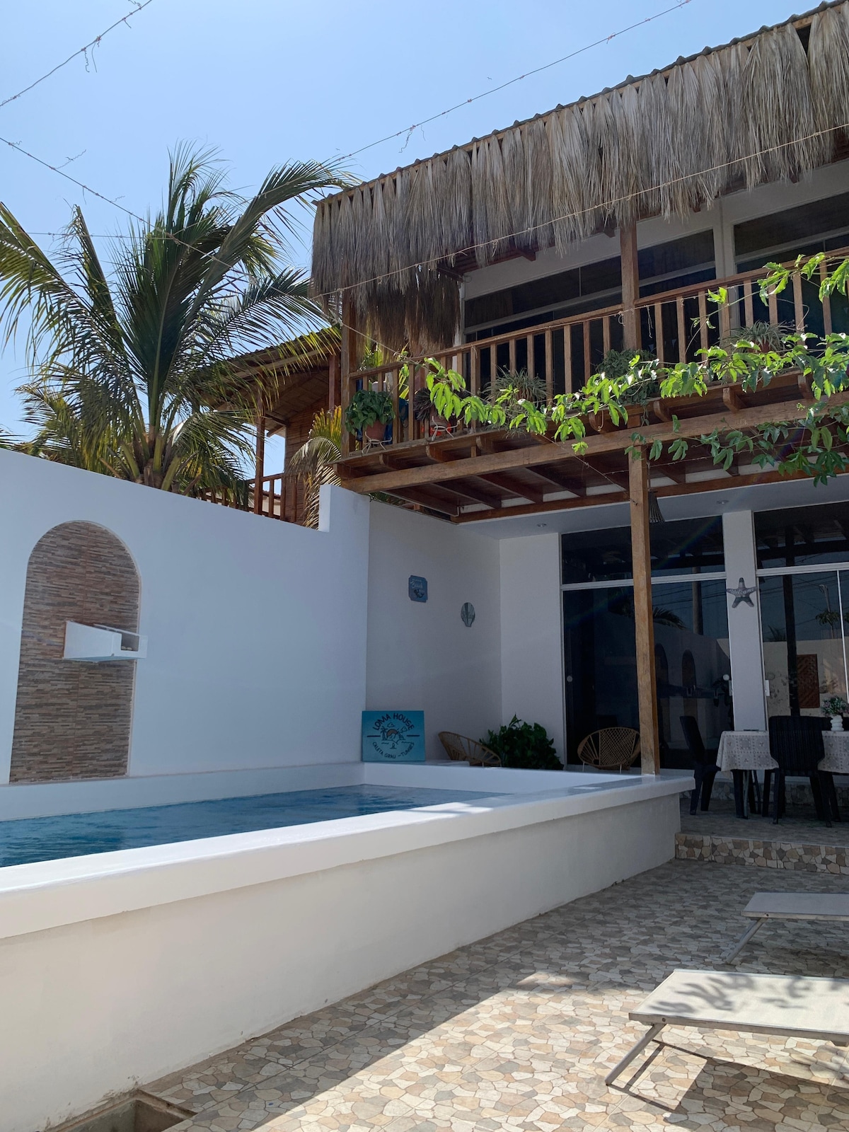 An outdoor pool is framed by a modern structure with tropical elements. The upper balcony is adorned with natural wood features and greenery. A tiled patio area includes several chairs, enhancing the space for relaxation and leisure.