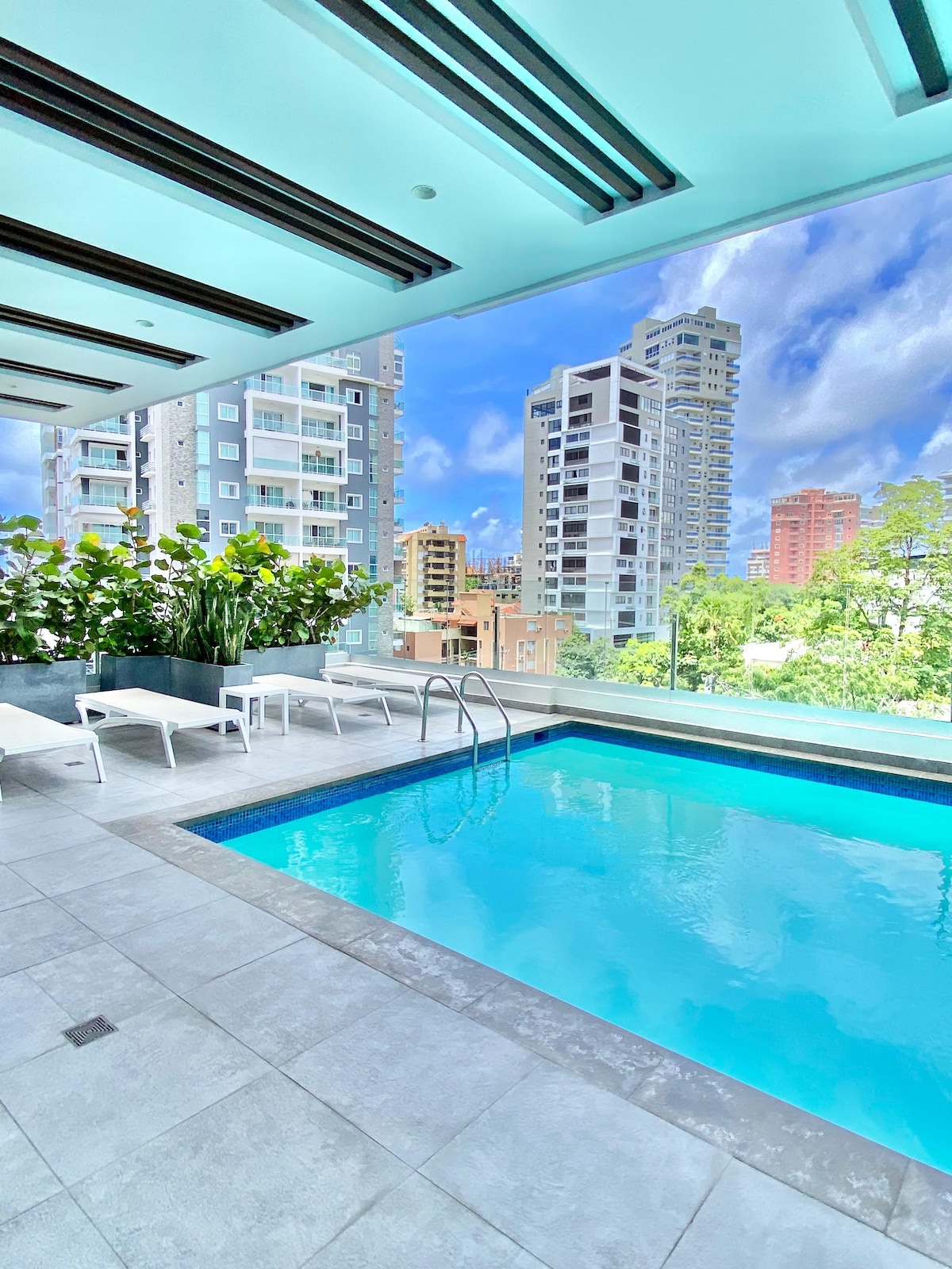 A modern pool area features a clear, inviting swimming pool surrounded by sleek tiles. Sun loungers are arranged beside the pool, with lush greenery adding a touch of nature. Panoramic windows offer a view of the cityscape and blue skies.
