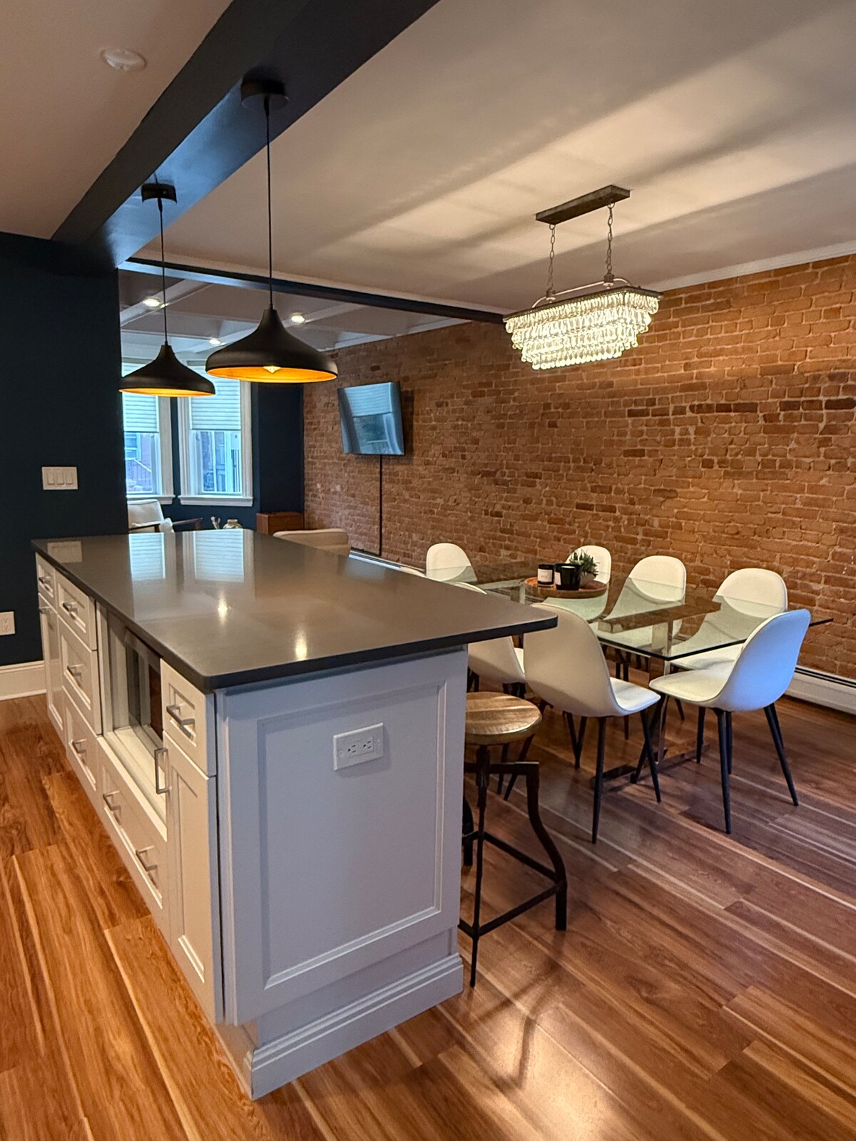 A modern kitchen area features a sleek island with bar seating and overhead pendant lights. A dining table for six is positioned adjacent to an accent brick wall, enhancing the space. Natural light enters through a window, illuminating the contemporary furnishings.