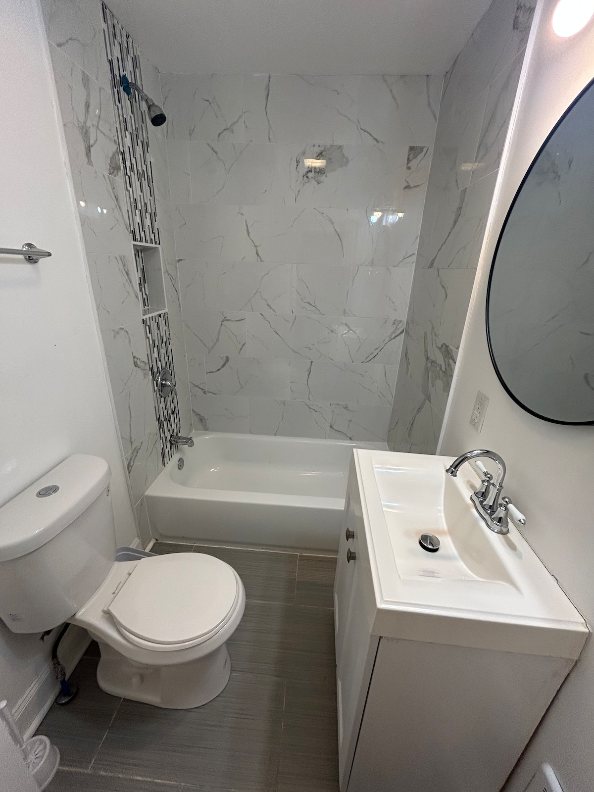 The bathroom features a modern design with marble-patterned walls and a sleek vanity. A bathtub is located adjacent to the shower area, while a round mirror above the sink adds a contemporary touch. Neutral colours create a clean and spacious appearance.