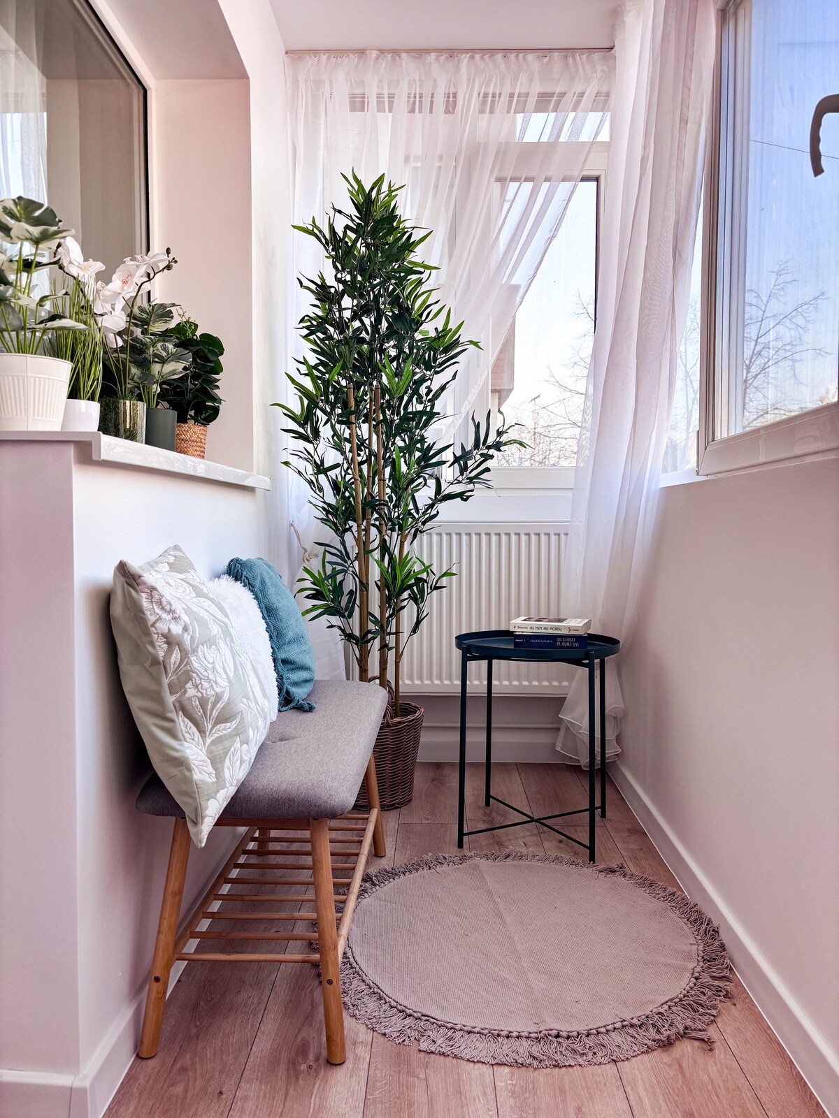 A cozy corner is highlighted by a bench adorned with decorative pillows. A small round table stands nearby, accommodating a stack of books. Bright green plants are positioned beside a large window, with sheer curtains allowing natural light to filter through.