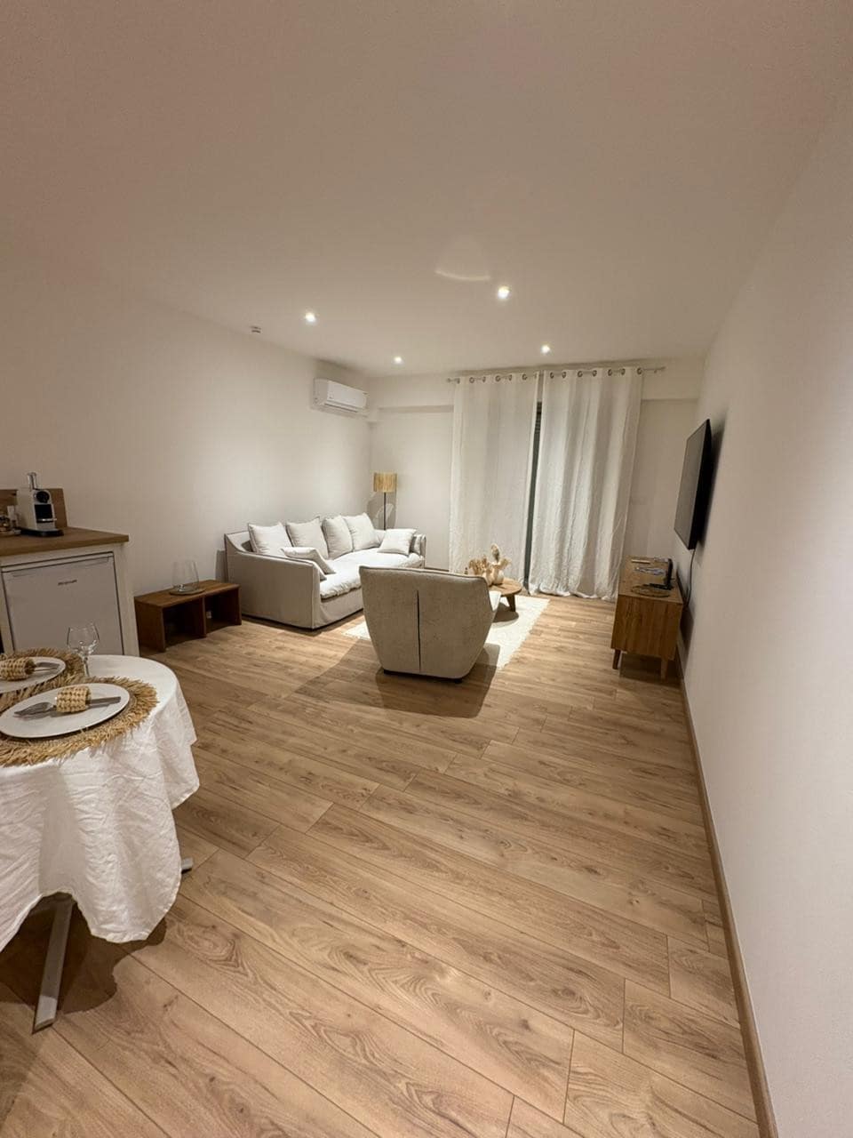 A spacious living area features a comfortable seating arrangement with a light-colored sofa and armchair. Soft lighting illuminates the wood-paneled floor, while white curtains frame a window. A small dining table is positioned nearby, set for a meal.