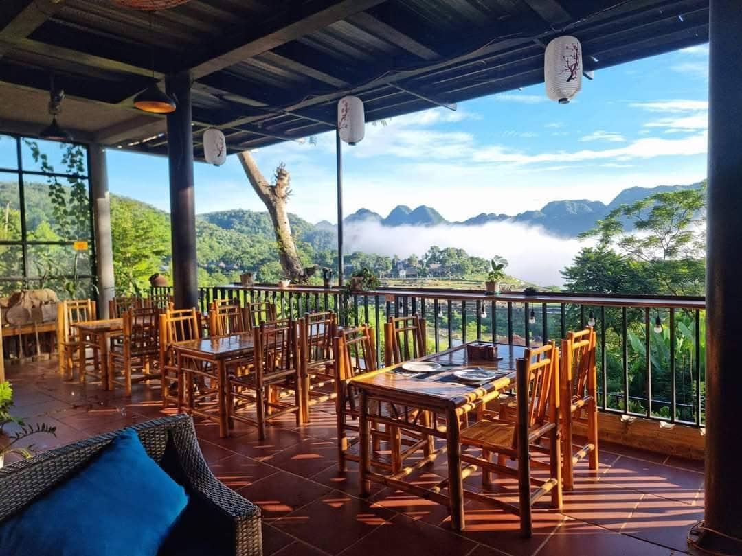 A spacious outdoor seating area features wooden tables and chairs arranged to provide views of the surrounding valley. Soft clouds are seen lingering in the landscape, with mountains rising in the background under a clear blue sky.