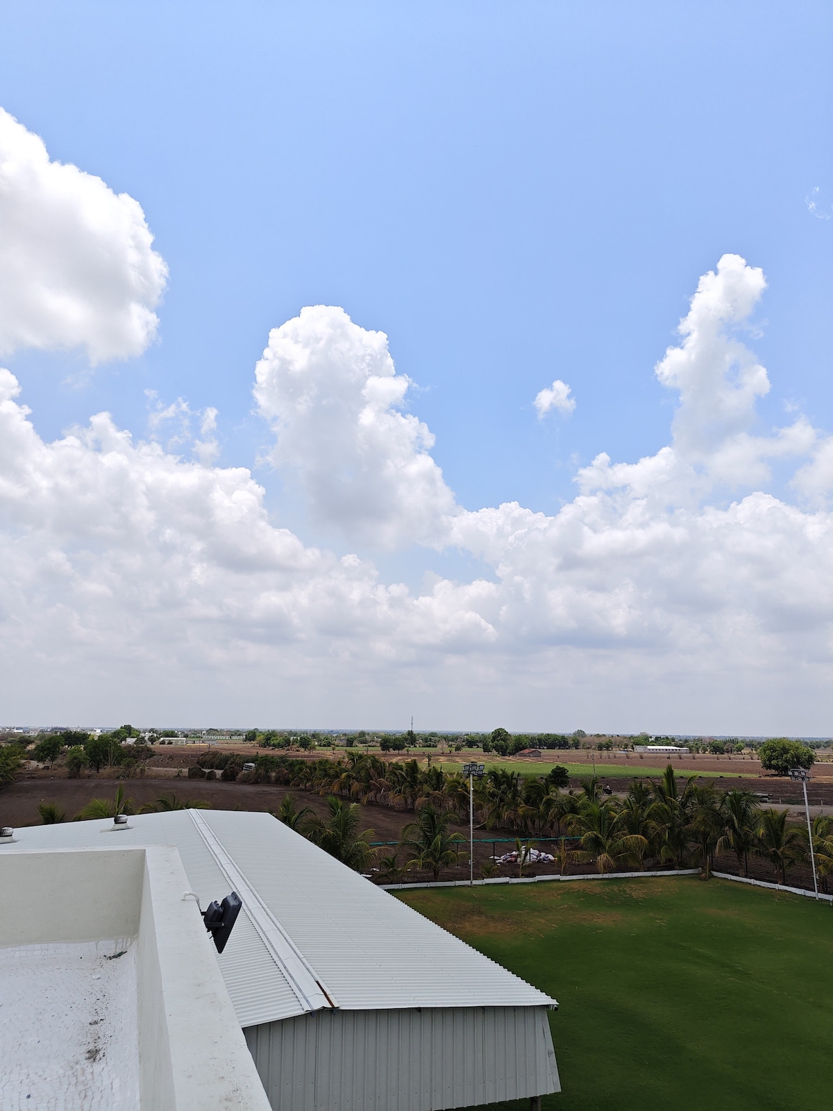 A panoramic view showcases a clear blue sky filled with scattered clouds above a lush green landscape. Expansive fields and trees can be seen in the distance, highlighting the tranquil atmosphere of the area.