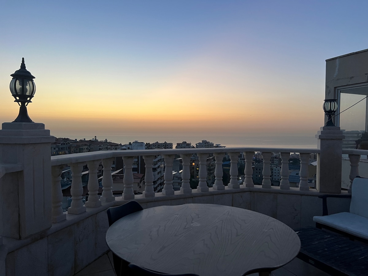 An exclusive terrace with a spacious table and outdoor seating captures a serene sunset over the sea. The balustrade framing the view features elegant lanterns, adding to the tranquil ambiance of the surrounding landscape.