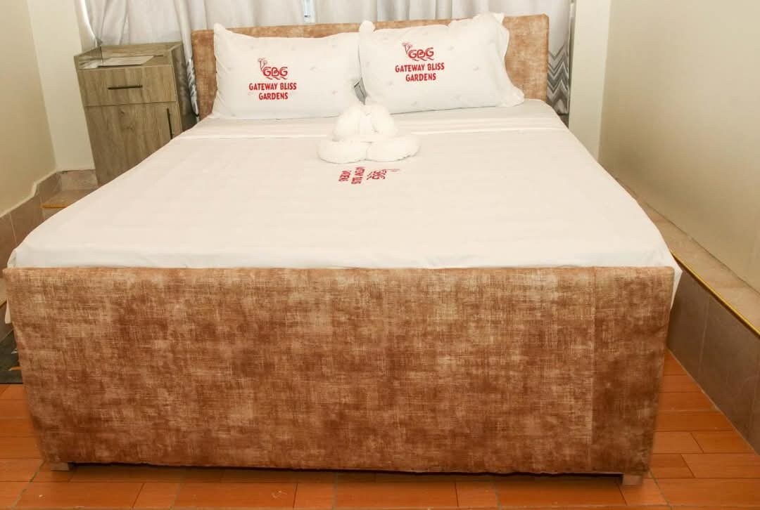 A spacious bed is centered in the room, featuring a tufted beige headboard and white linens. Two decorative pillows are placed at the head of the bed, along with a neatly arranged towel shaped like a swan. A bedside table is positioned next to the bed.