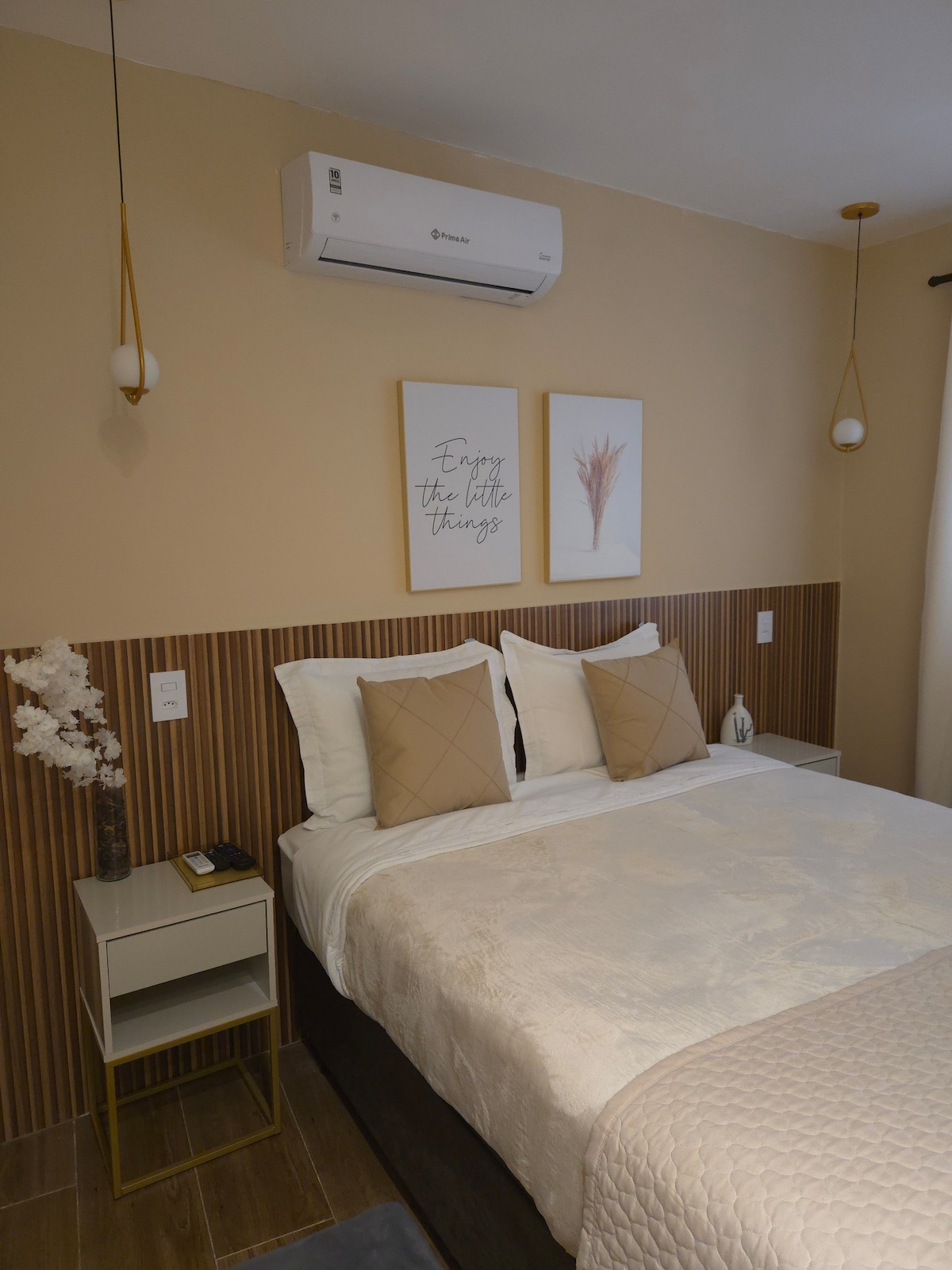 A spacious bedroom is equipped with a large bed featuring neutral-toned bedding and decorative pillows. The wall behind the bed is adorned with wooden panels and framed art pieces. Soft lighting is provided by pendant lamps, while a small bedside table holds a phone and a decorative element.