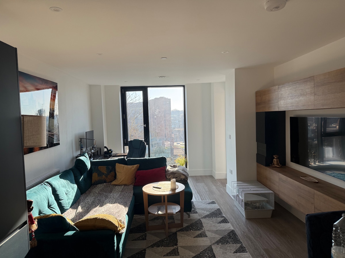 A modern living area features a bright green sofa adorned with a textured throw and decorative cushion. A wooden coffee table sits in front, accompanied by a patterned rug. Large windows allow natural light to illuminate the space, offering views of the city.