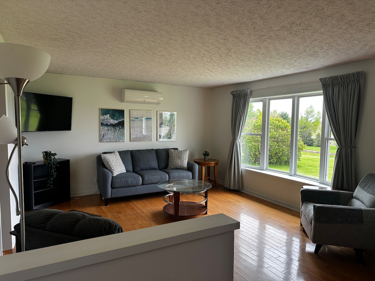 A spacious living area features a comfortable couch and armchair, arranged around a glass coffee table. Natural light filters through large windows, illuminating artwork on the walls. A flat-screen television is mounted opposite the seating, and greenery is visible outside.