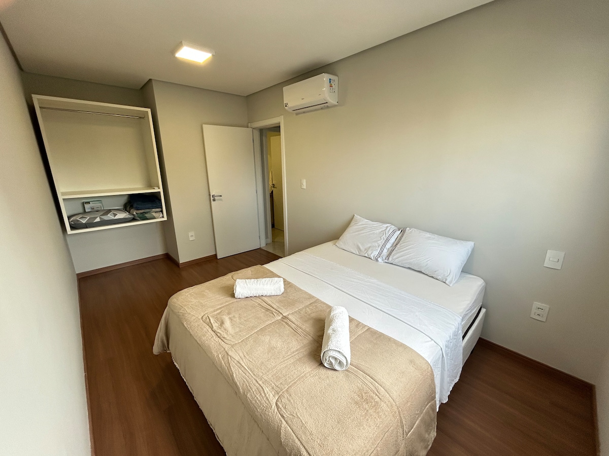 A neutral-toned bedroom features a spacious bed with a light blanket and two rolled towels placed at the foot. A built-in closet offers storage space, and a minimalist decor enhances the room's functionality. Natural light enters through a nearby doorway.