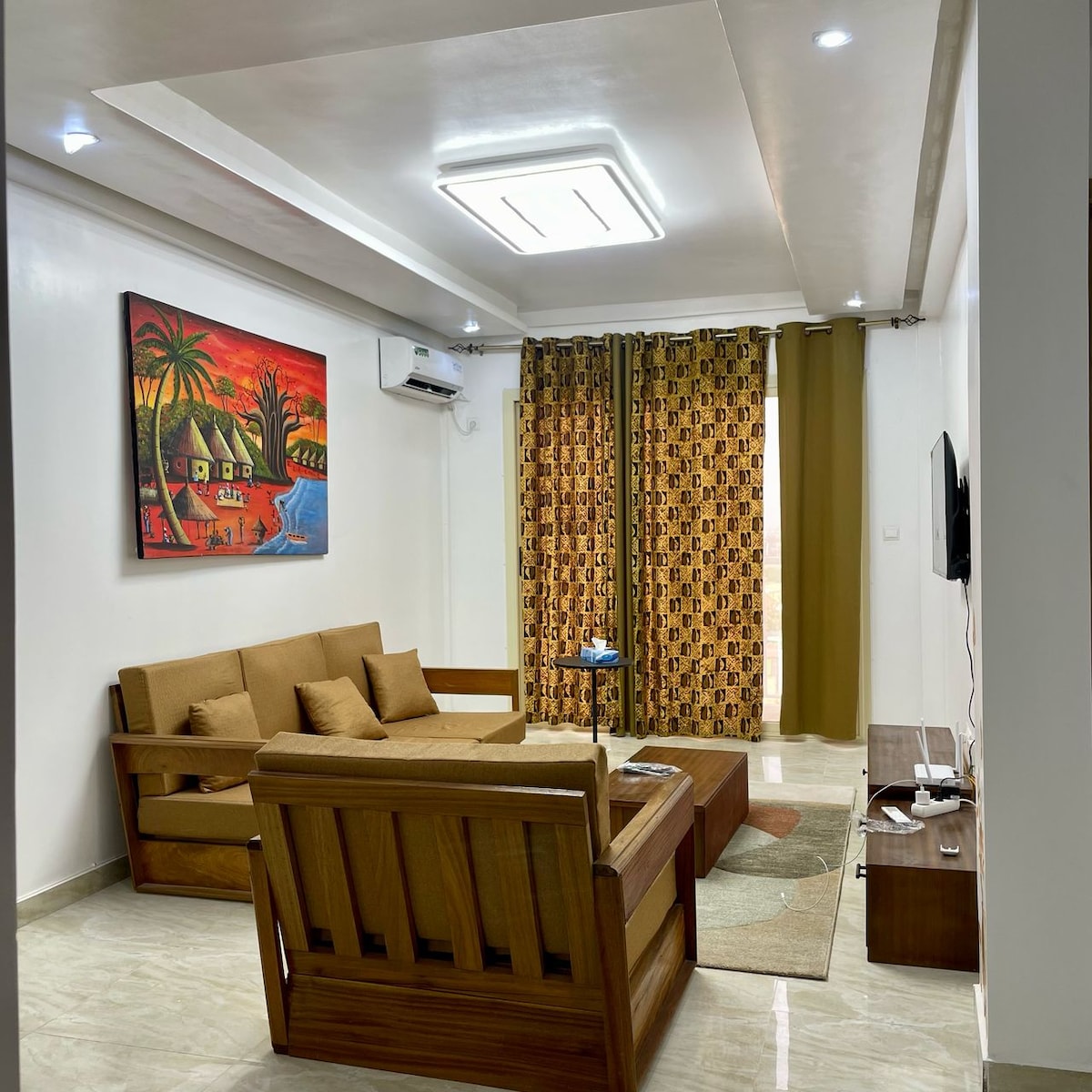 A living room area is furnished with a spacious sofa set and a wooden coffee table. Large curtains with a patterned design frame the windows, while colorful wall art adds a vibrant touch. A television is mounted on the wall, complementing the modern decor.