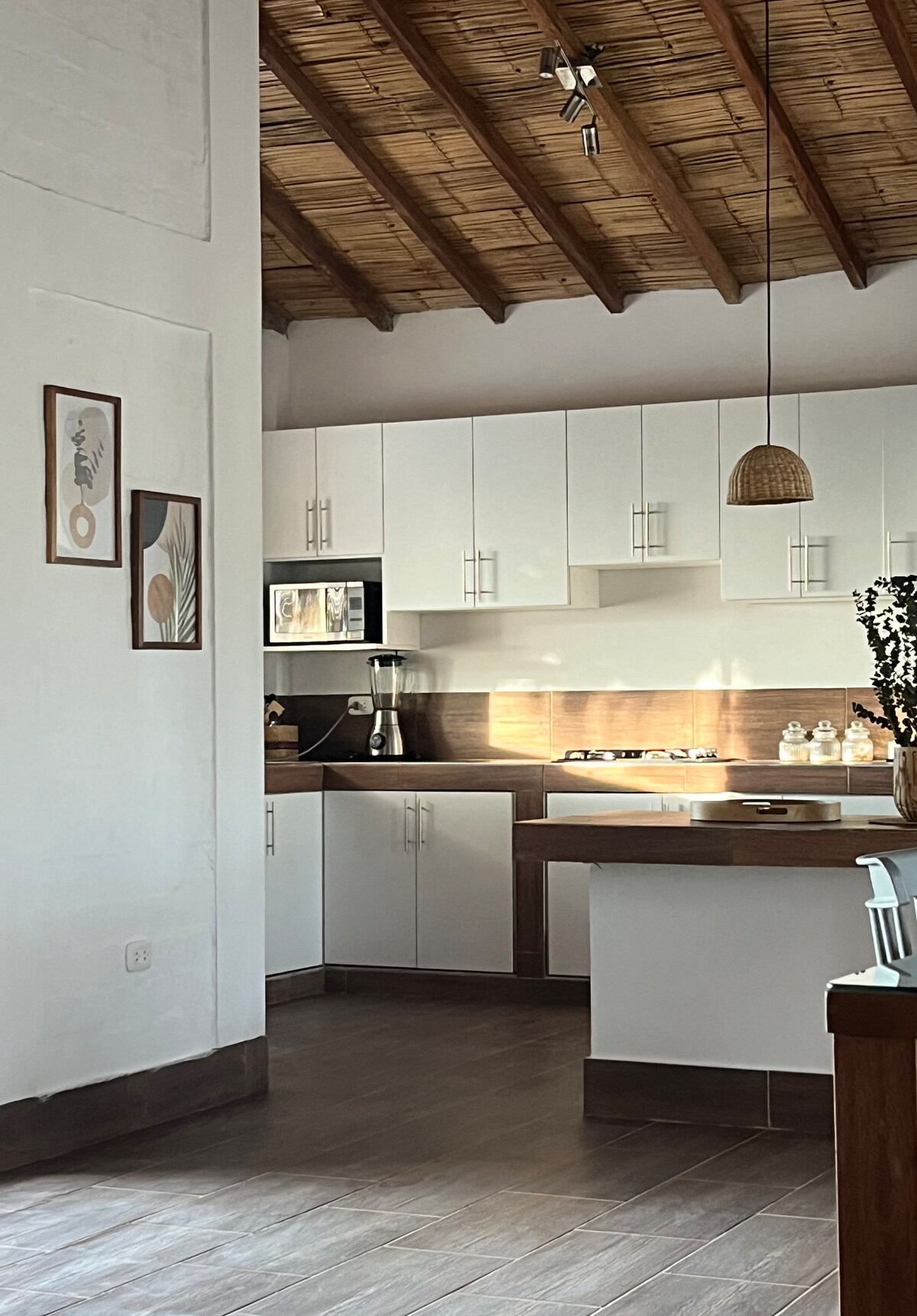 The modern kitchen features crisp white cabinetry and a wooden countertop. A microwave and coffee maker are visible, along with a gas stove. Natural light fills the space, highlighting decorative elements like framed art and a woven pendant light hanging from the thatched ceiling.
