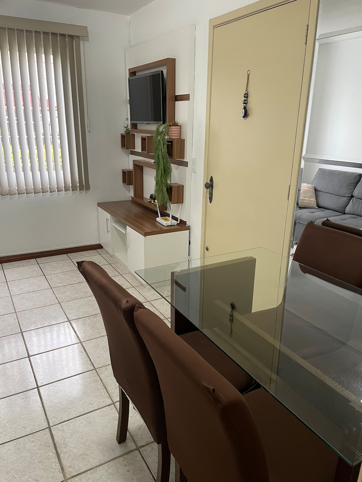 A dining area is displayed with a glass table surrounded by four brown chairs. A simple wall-mounted entertainment unit features a TV and decorative shelves. Vertical blinds allow natural light to filter in, illuminating the tiled floor.