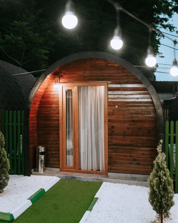 An inviting entrance to a uniquely shaped wooden structure is highlighted by soft lighting from overhead bulbs. A neatly trimmed green pathway leads to the door, flanked by shrubs and gravel, creating a welcoming approach to the distinctive lodging.