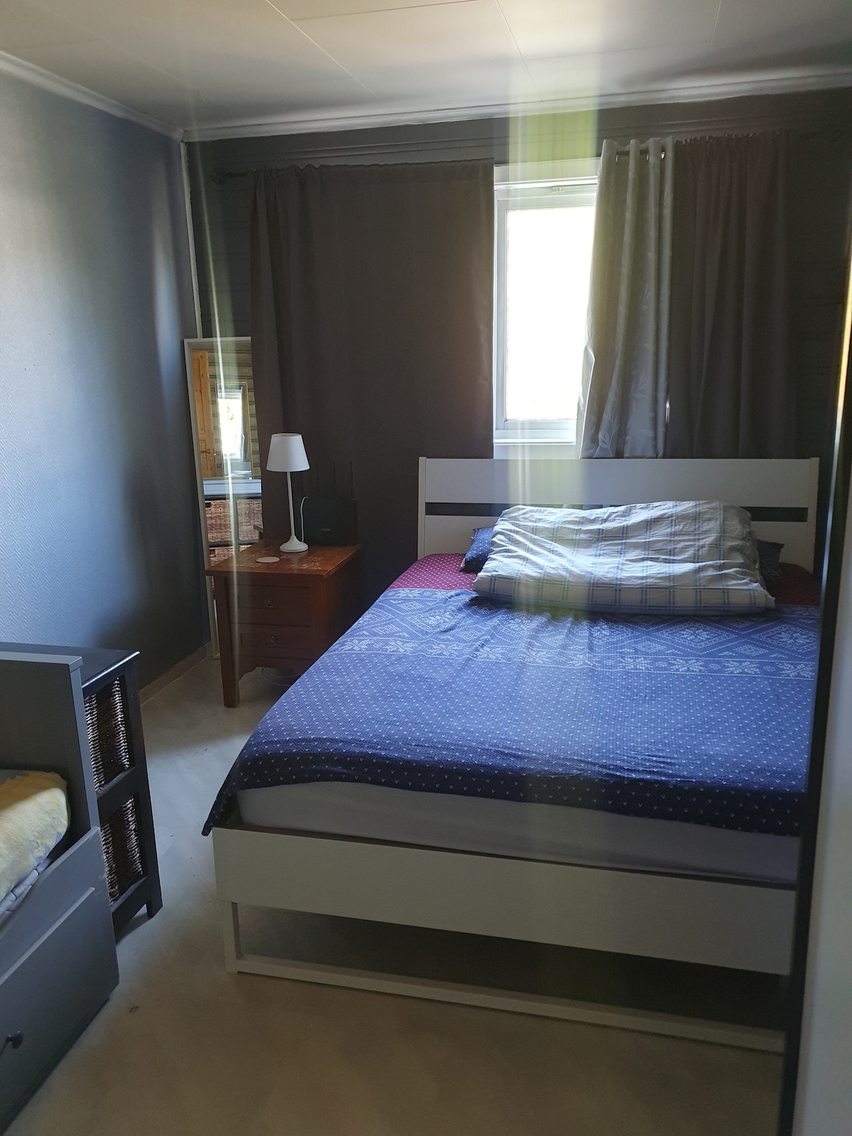 A cozy guestroom features a spacious bed with a patterned duvet cover, complemented by a bedside lamp and wooden nightstand. Natural light enters through a window, partially covered by dark curtains. A wardrobe and a mirror are also present, enhancing the room's functionality.