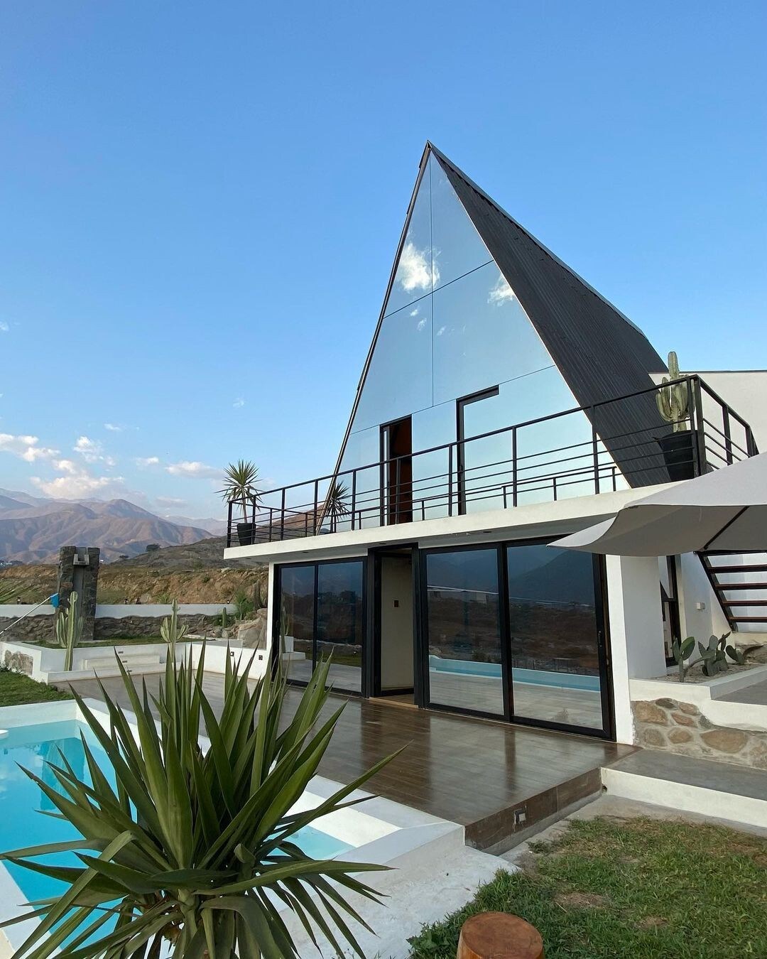 A unique architectural structure is displayed, featuring a striking triangular roof and large glass windows. The outdoor area includes a swimming pool, surrounded by greenery. The mountainous backdrop enhances the serene setting, and a wooden deck provides access to the interior.