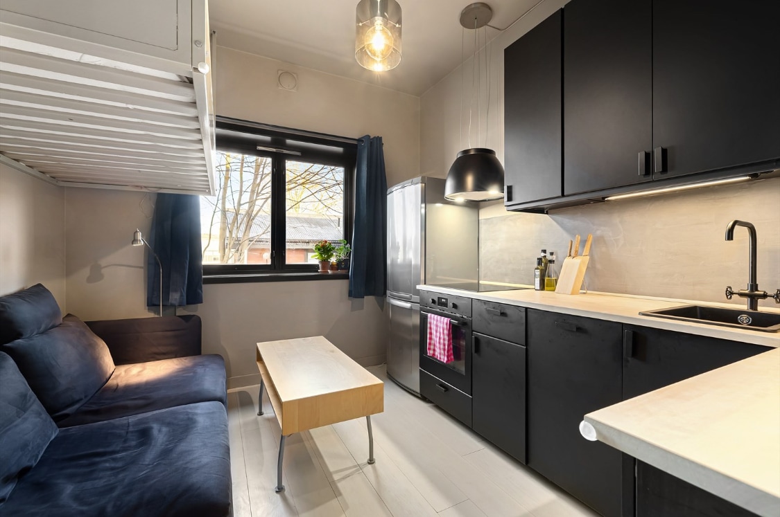 A well-equipped kitchen is complemented by a comfortable seating area. Light-colored cabinetry contrasts with darker elements, while a spacious countertop provides ample prep space. A compact living area features a sofa and a wooden coffee table. Large windows allow natural light to enhance the room.