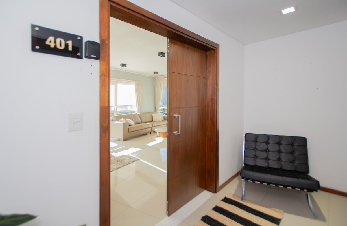 The entrance to apartment 401 is showcased with a modern wooden door. A black lounge chair is positioned beside the entryway, offering a seating option. Natural light is observed in the distance, illuminating a spacious living area visible through the doorway.